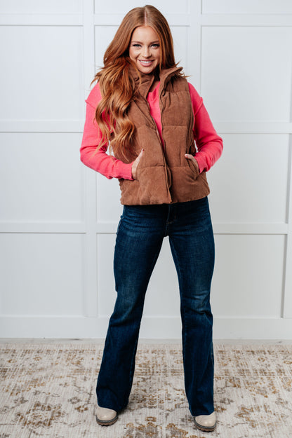 Woman wearing a brown vest over a pink shirt with blue jeans against a white paneled wall.