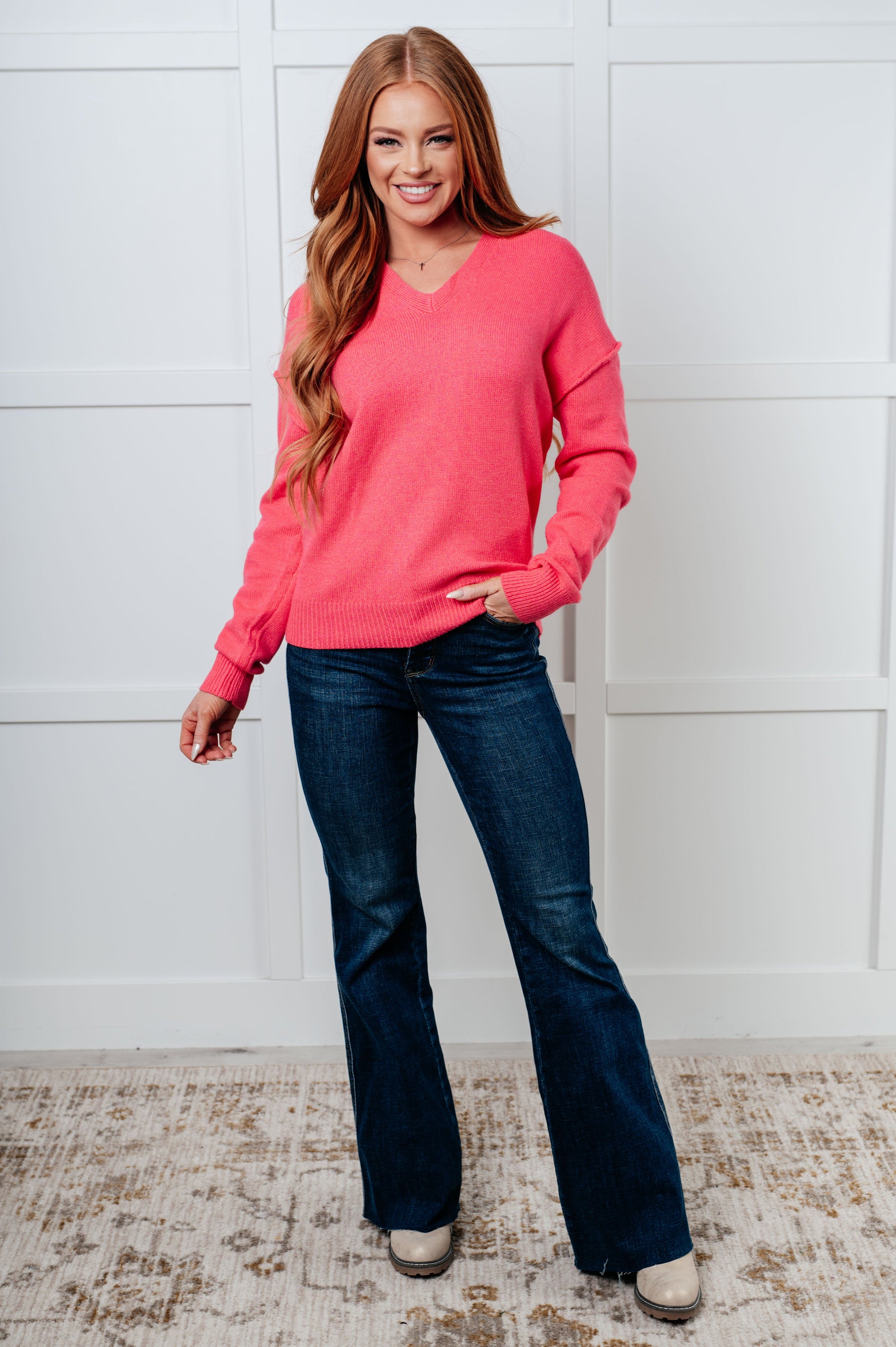 Woman wearing a pink sweater and blue jeans standing against a white wall.