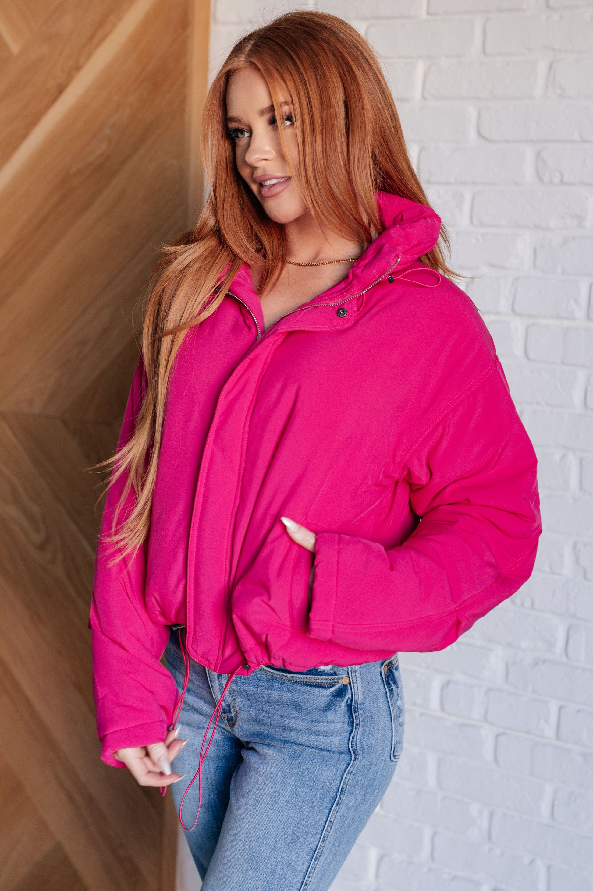 Woman wearing a bright pink puffer jacket against a white brick wall.