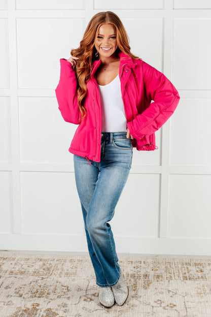 Woman wearing a bright pink jacket, white top, and blue jeans against a white tiled wall.