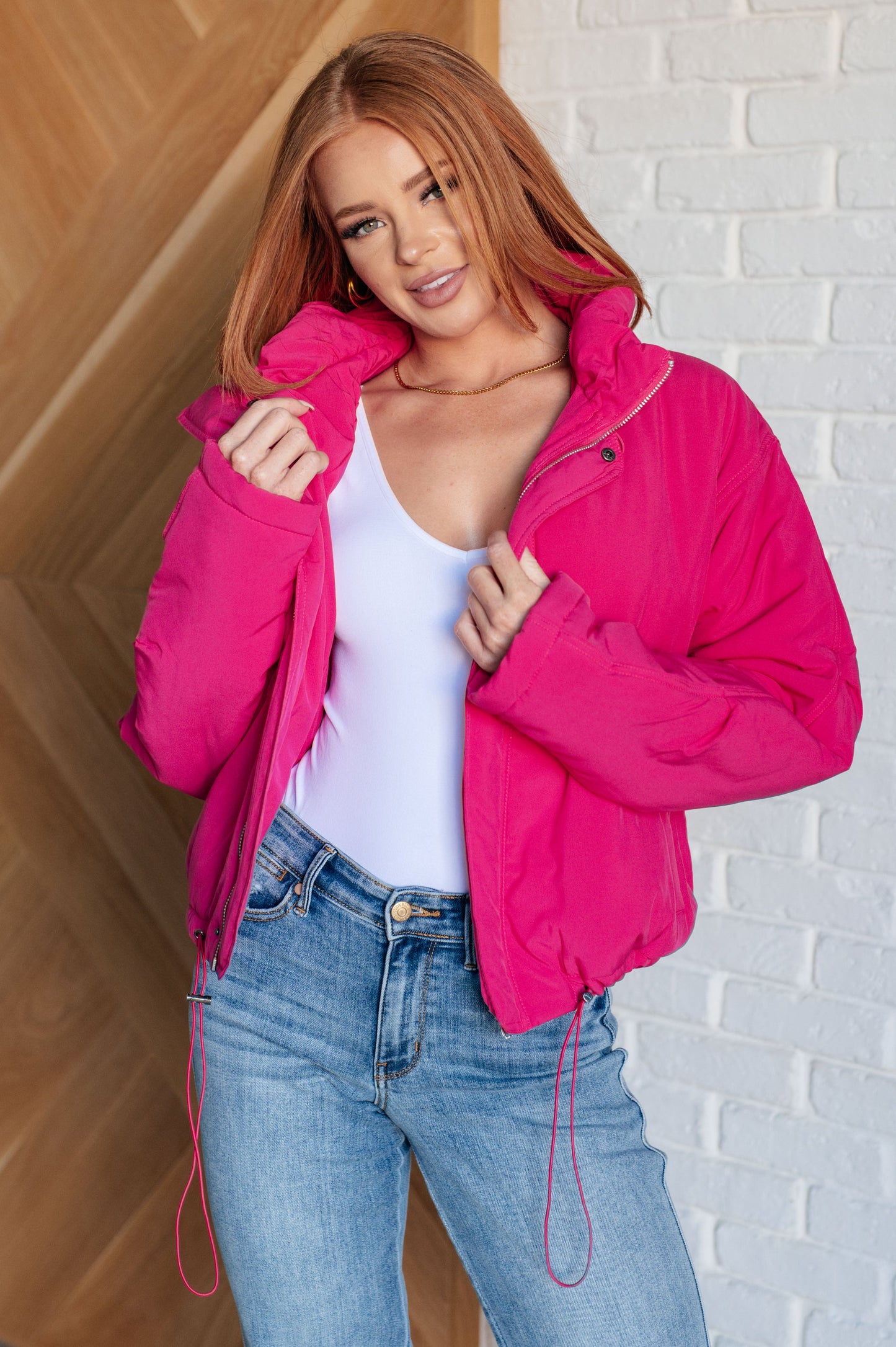 Woman wearing a bright pink jacket over a white top and blue jeans against a white brick wall.