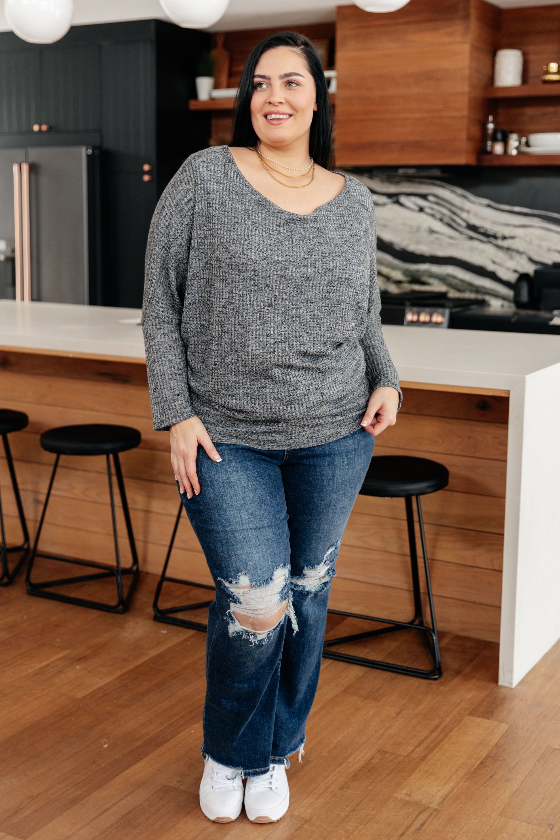 Woman standing in a kitchen wearing a gray sweater and blue jeans.