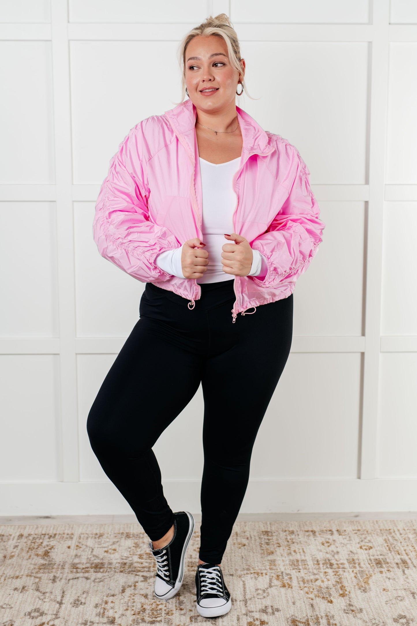 Person wearing a pink jacket, white shirt, black pants, and black sneakers against a white wall.
