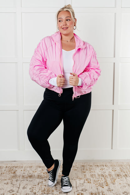 Person wearing a pink jacket, white shirt, black pants, and black sneakers against a white wall.
