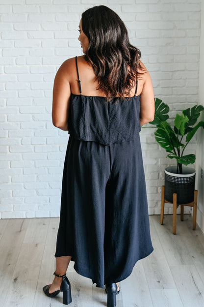Woman wearing a black jumpsuit standing in a room with a white brick wall and a plant.