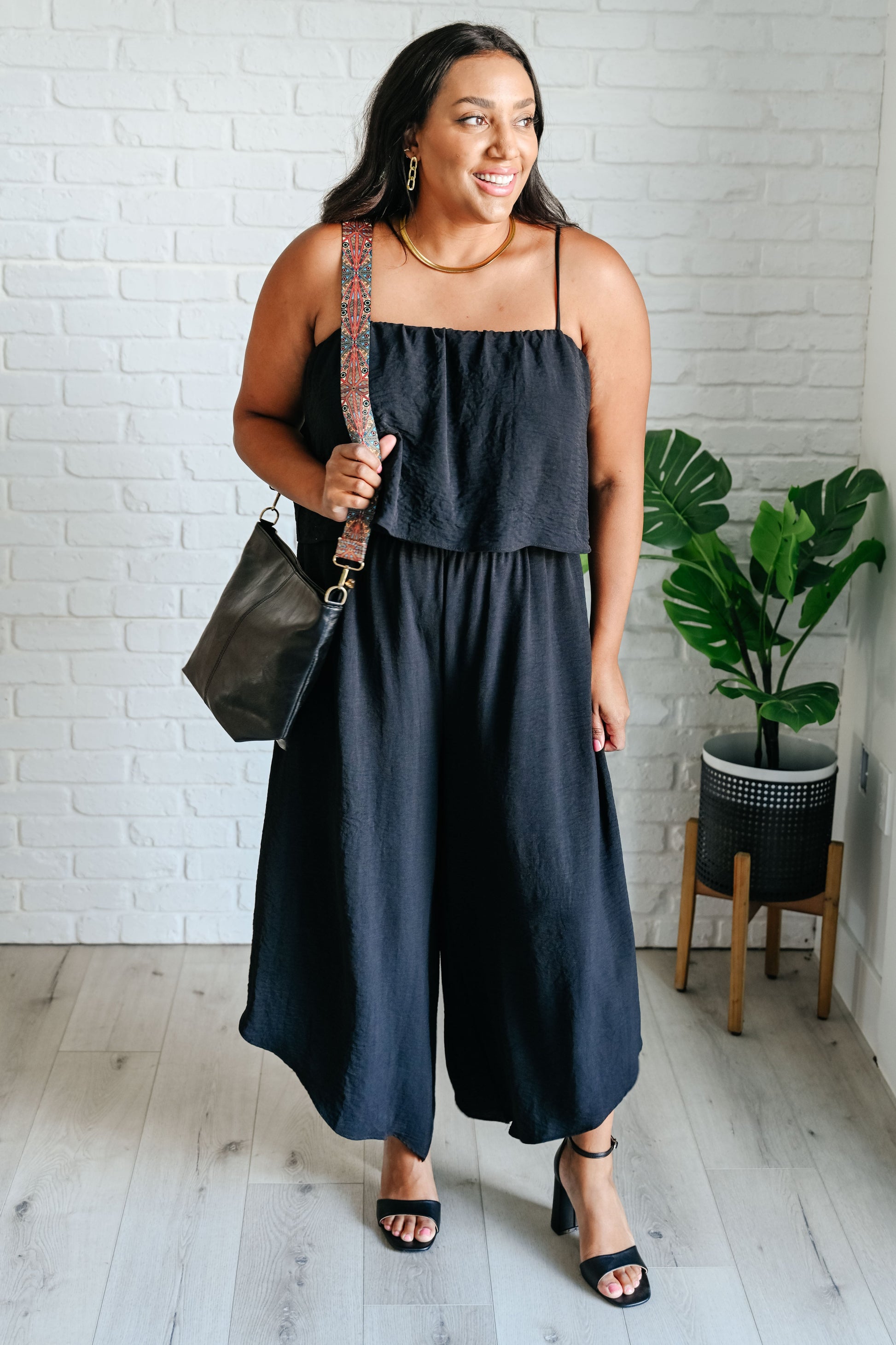 Woman wearing a black jumpsuit holding a black bag in a room with a white brick wall and plant.