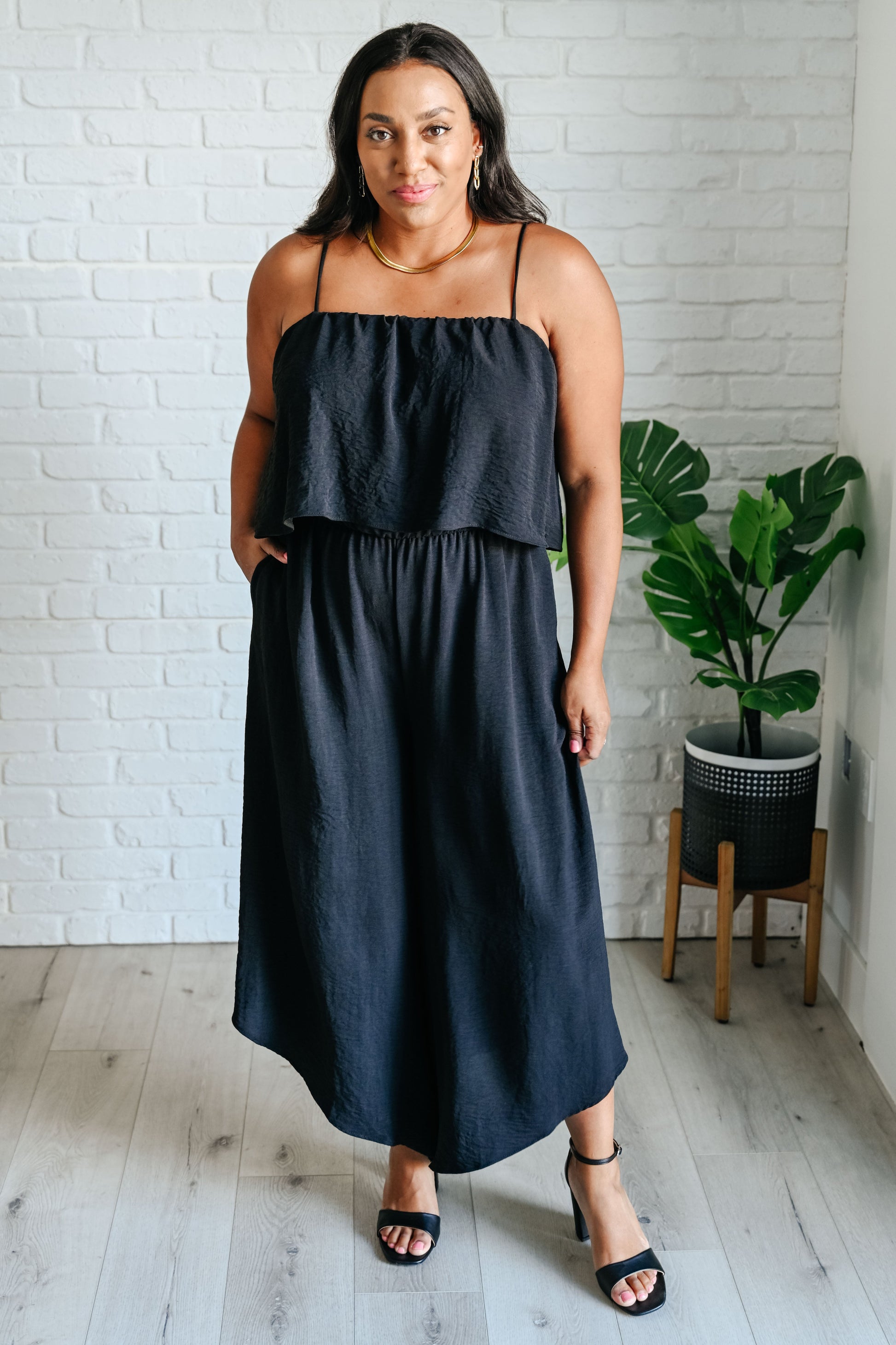 Woman wearing a black dress standing in a room with a white brick wall and a plant.
