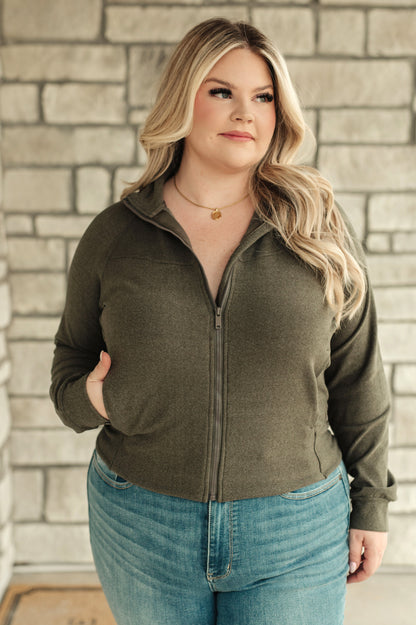 Woman wearing an olive green zip-up sweater and blue jeans against a brick wall.