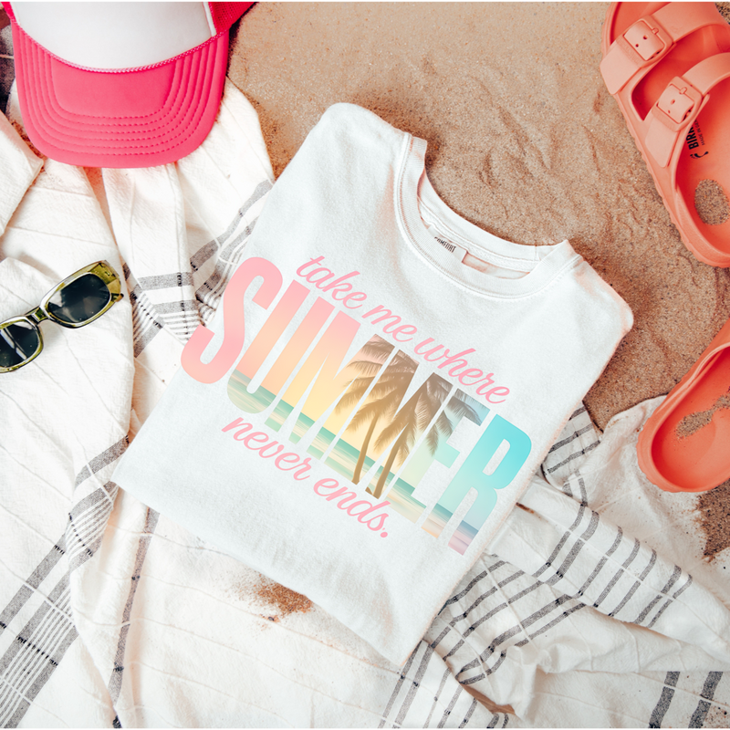 White t-shirt with colorful text on a sandy surface with beach accessories.