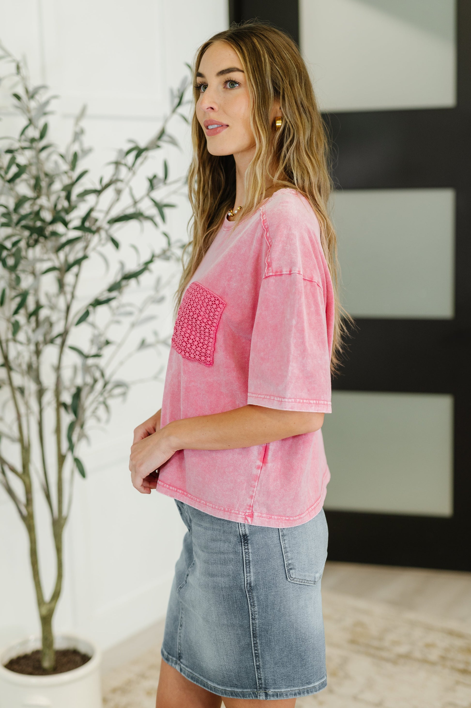 Woman wearing a pink shirt with a pocket detail and denim skirt indoors.