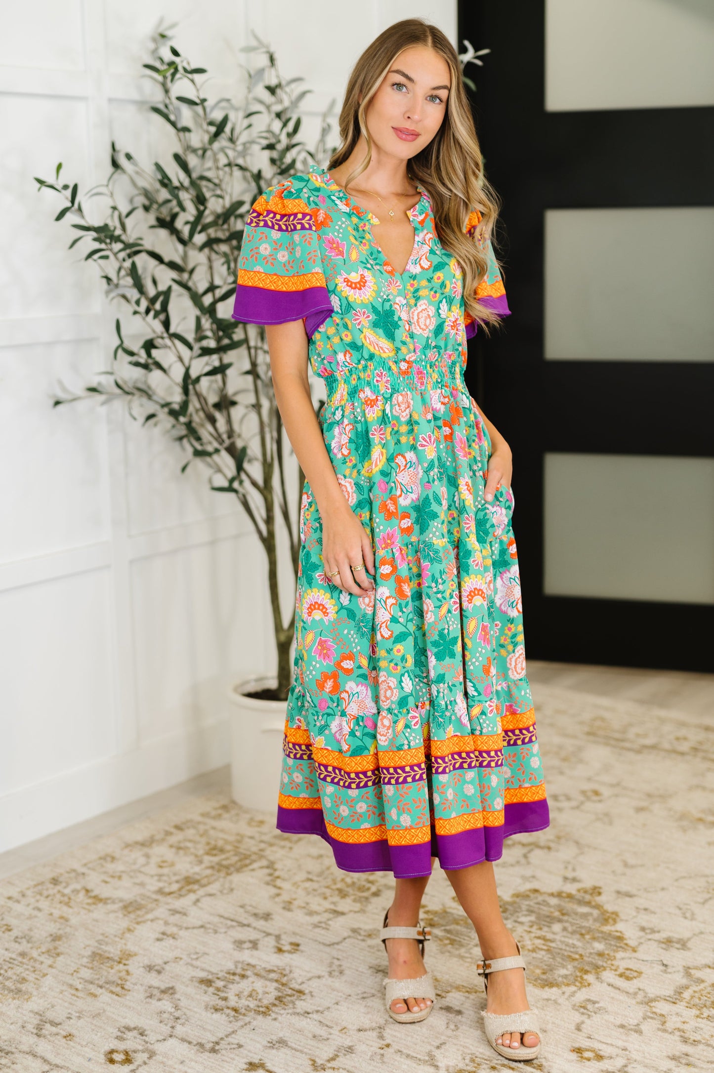 Woman wearing a colorful floral dress standing indoors with a plant in the background