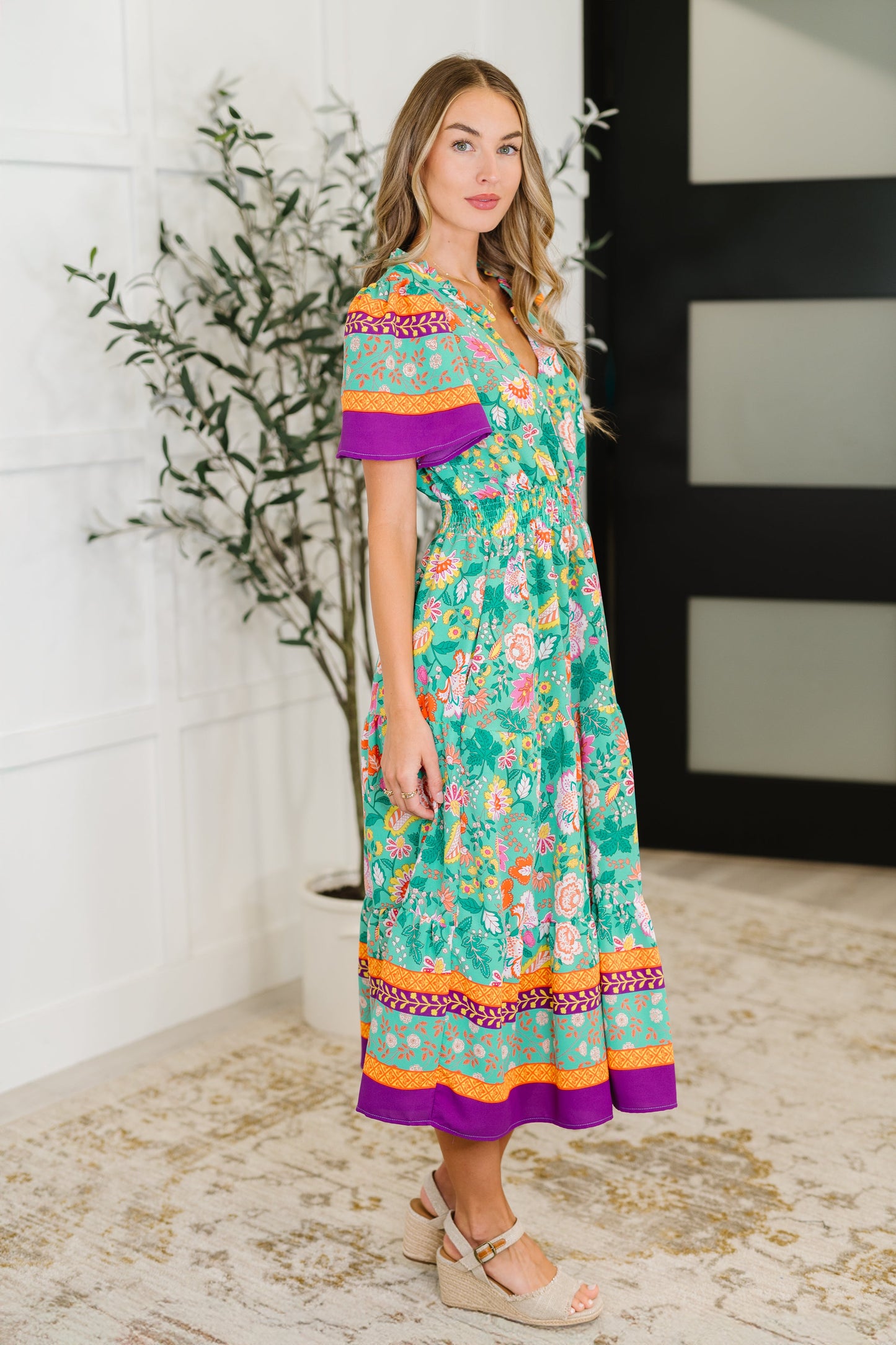 Woman wearing a colorful floral dress with a white wall and plant in the background