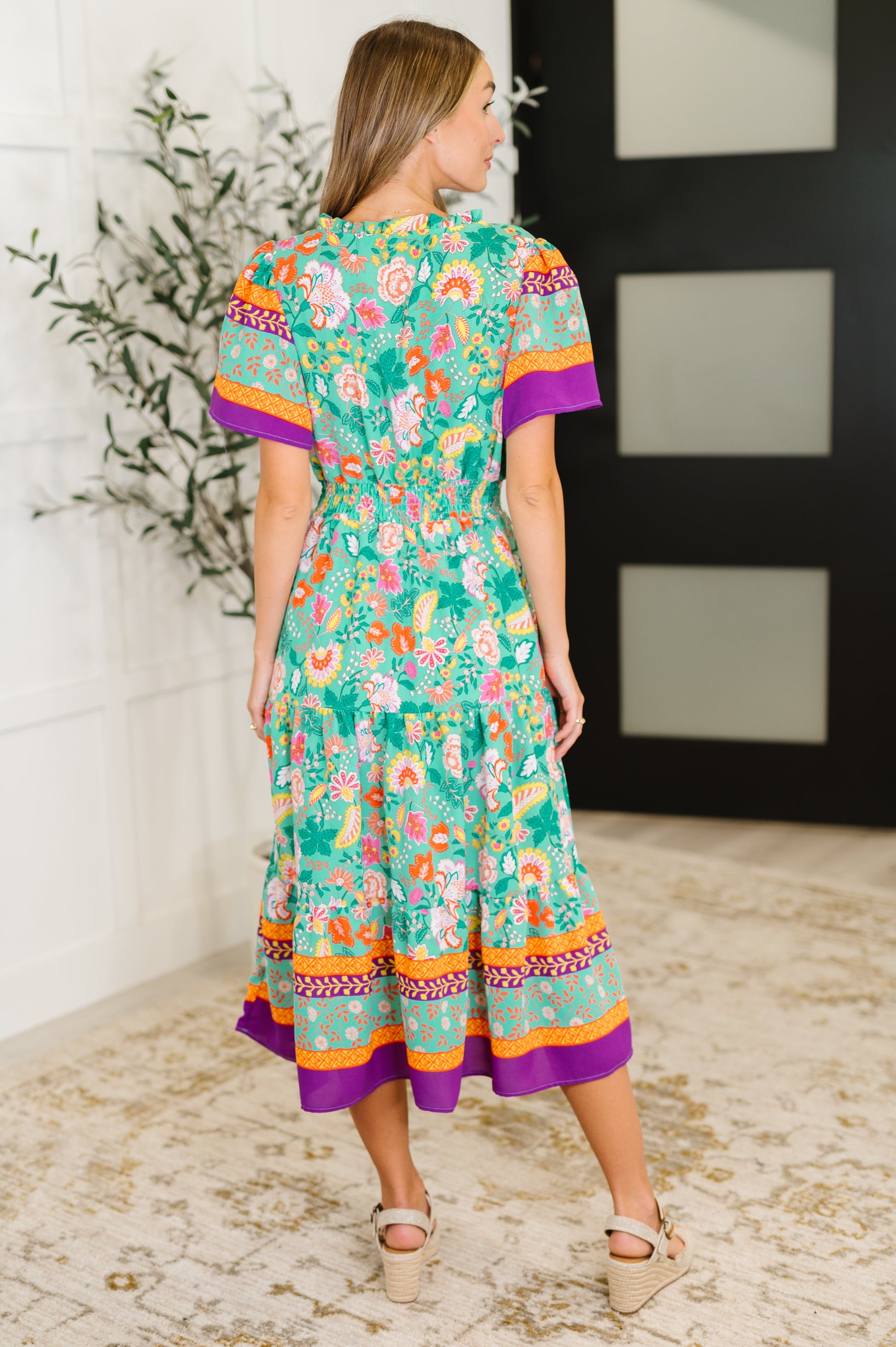Woman wearing a colorful floral dress standing in a room with a white wall and plant.