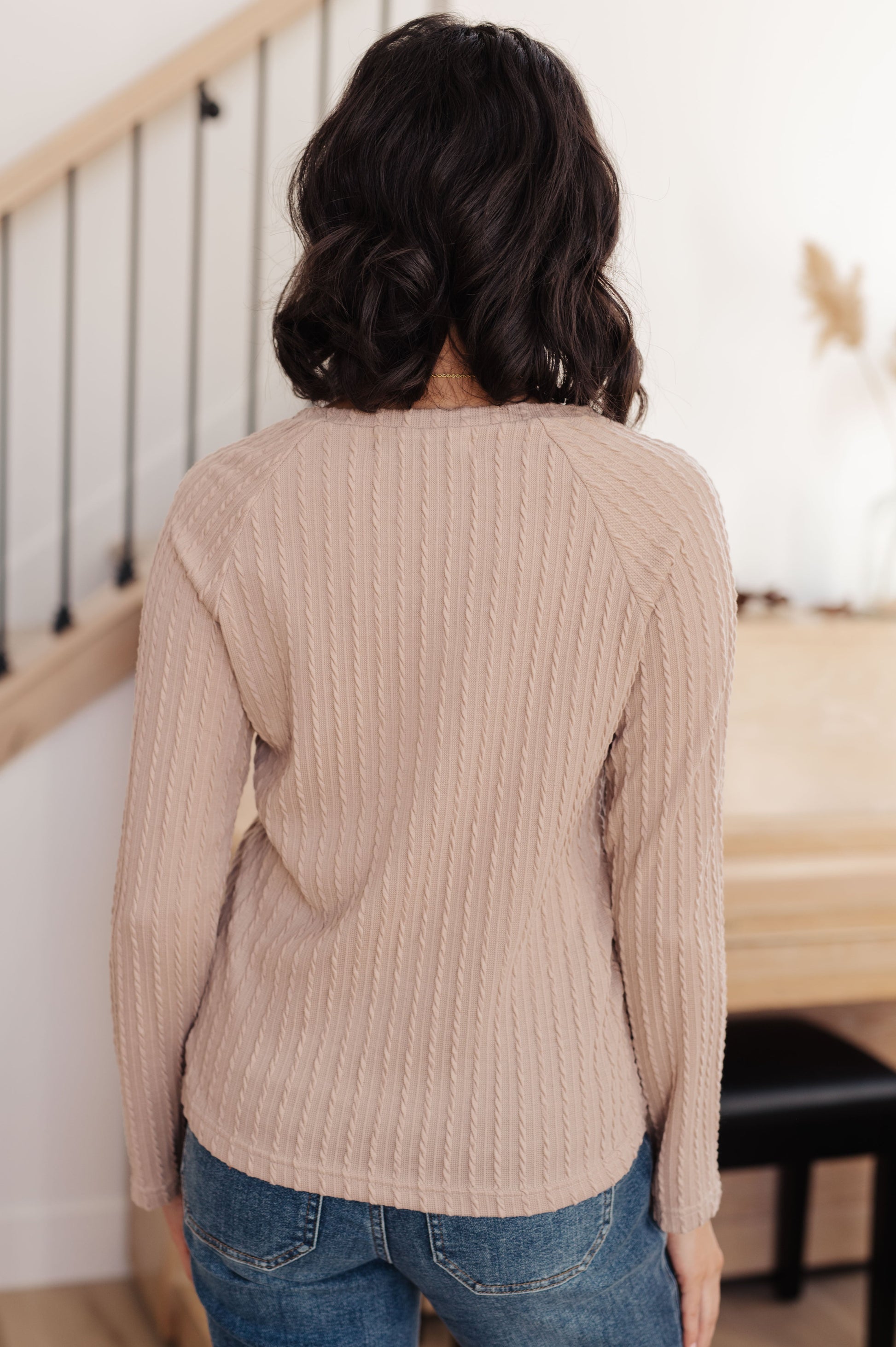 Person wearing a beige sweater and blue jeans standing in a room with a staircase.