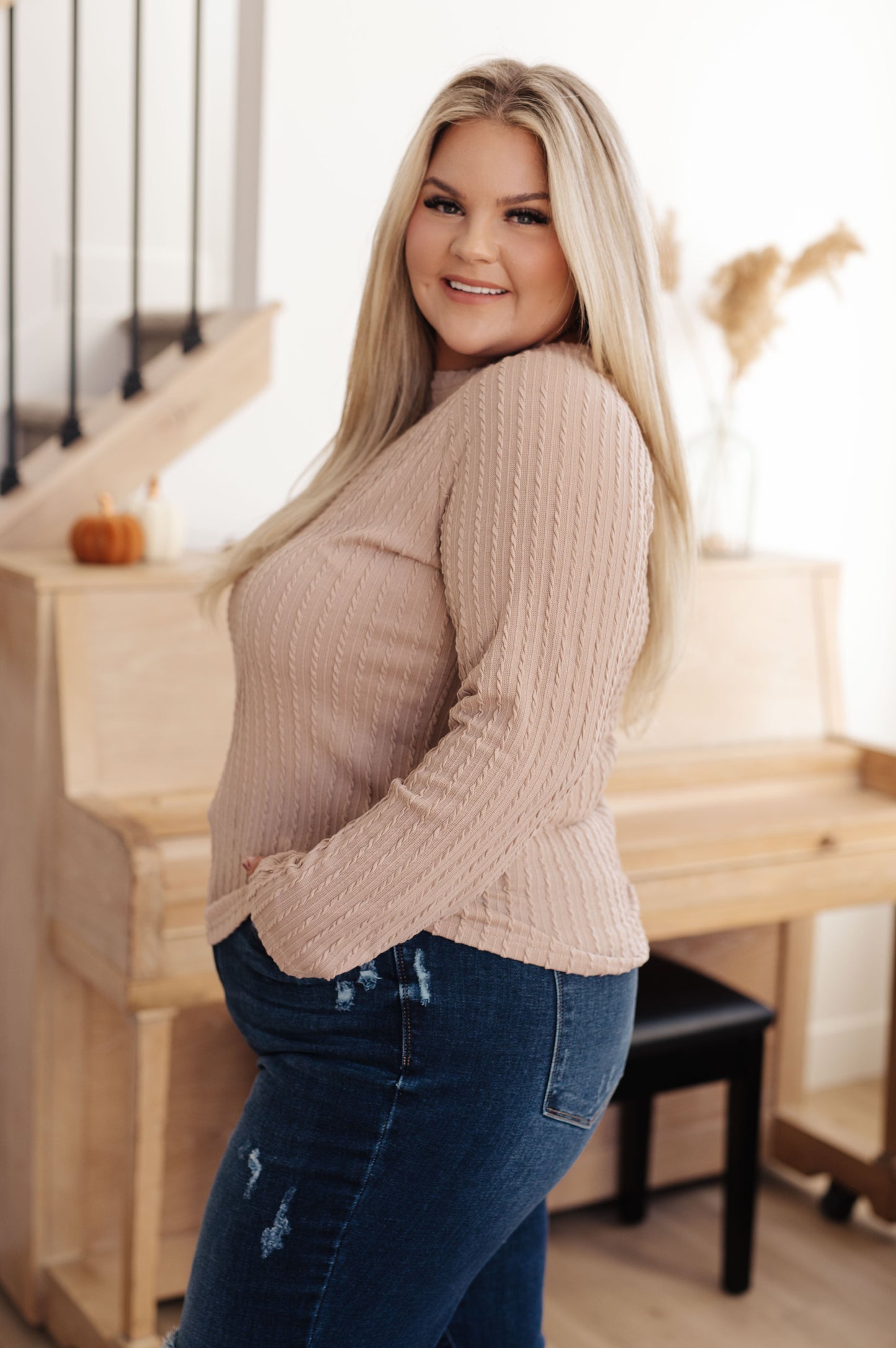 Woman wearing a beige sweater and blue jeans standing in a home setting.