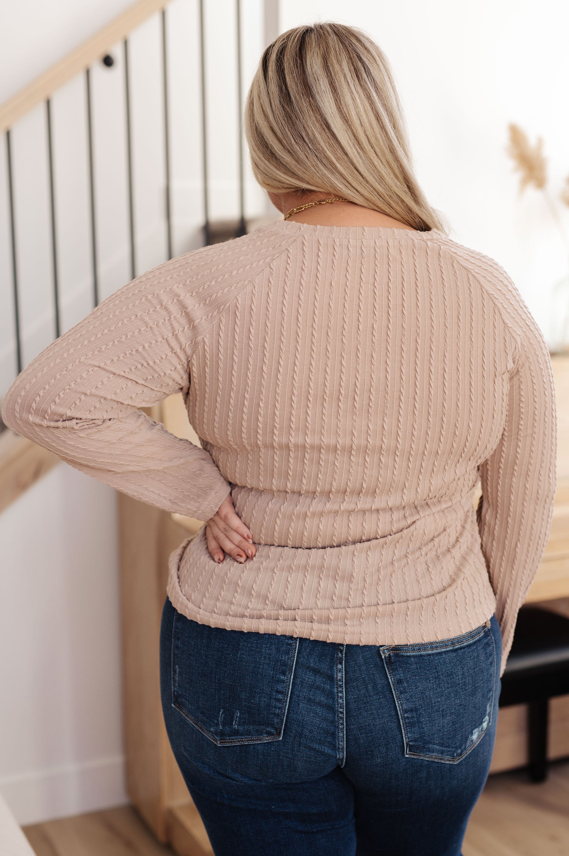 Person wearing a beige ribbed sweater and blue jeans standing in a room with wooden flooring and a white wall.