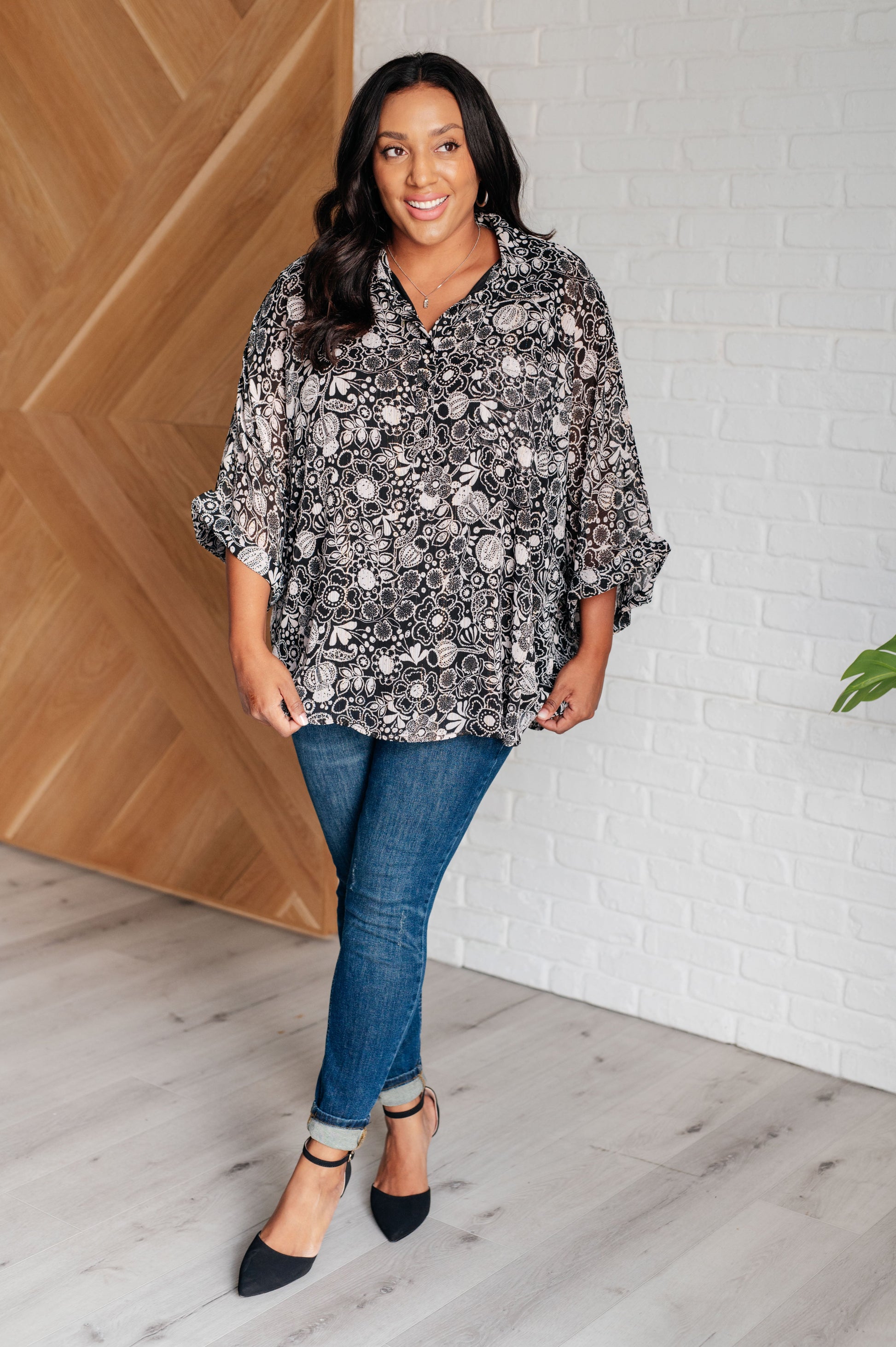 Woman wearing a floral blouse and jeans standing against a white brick wall.