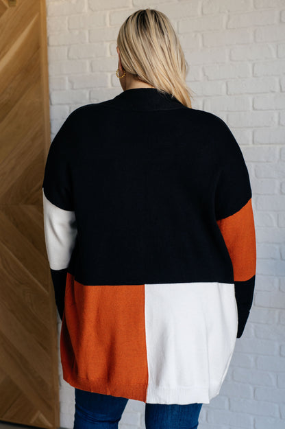 Person wearing a black, orange, and white color-blocked sweater against a white brick wall.