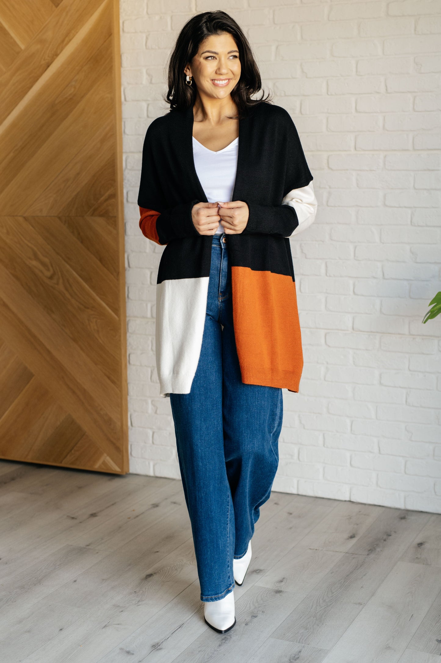 Woman wearing a black, orange, and white cardigan with blue jeans indoors.