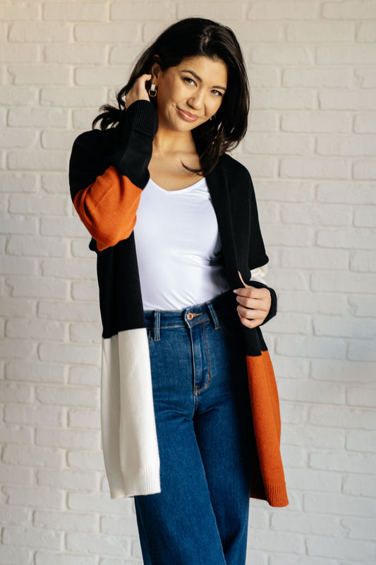Woman wearing a colorful cardigan over a white top and blue jeans, holding an orange bag against a white brick wall.