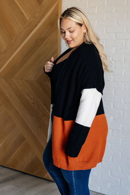 Woman wearing a black, white, and orange color-blocked sweater against a wooden and white brick wall.