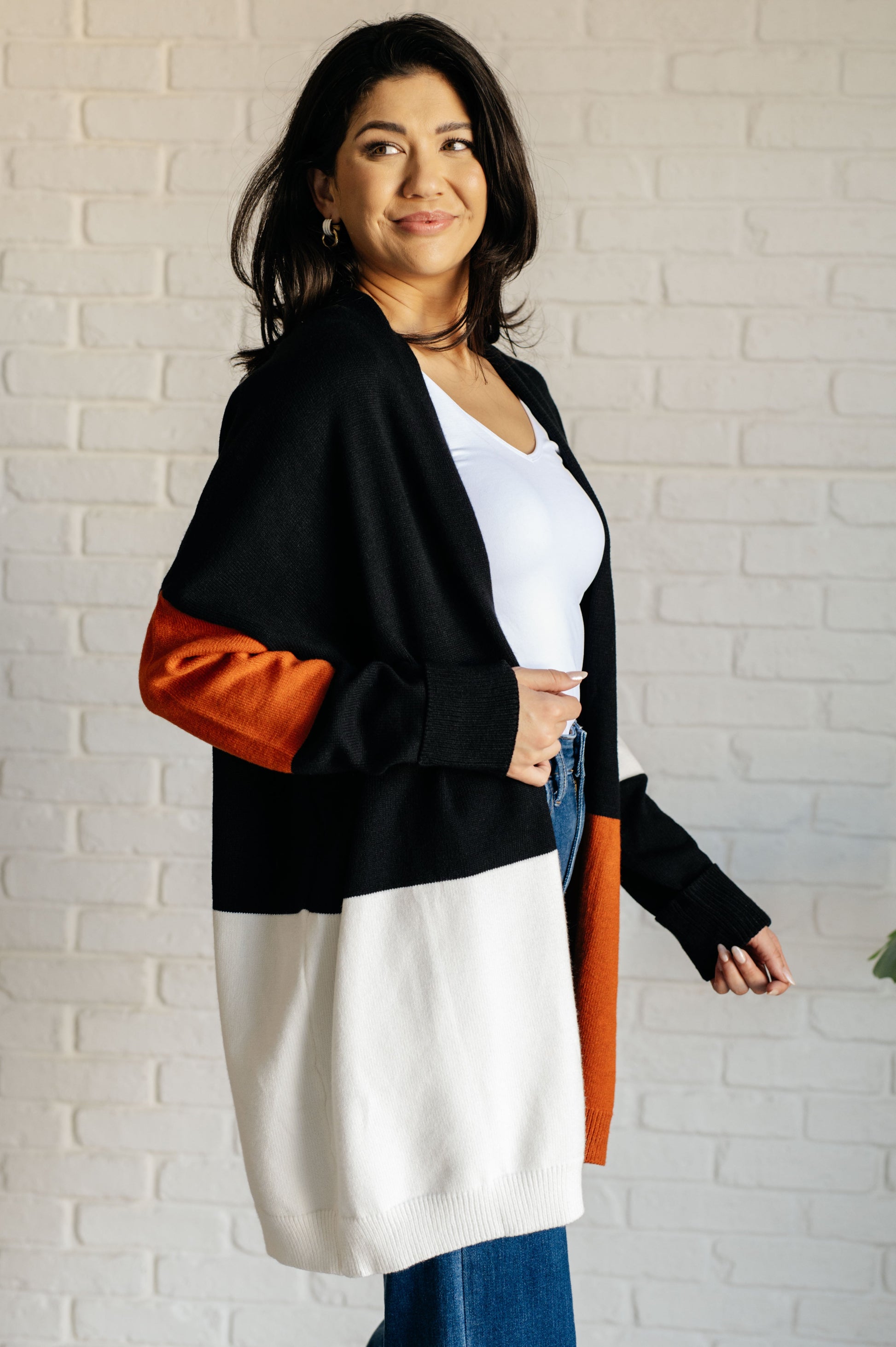 Woman wearing a black, orange, and white cardigan against a white brick wall.