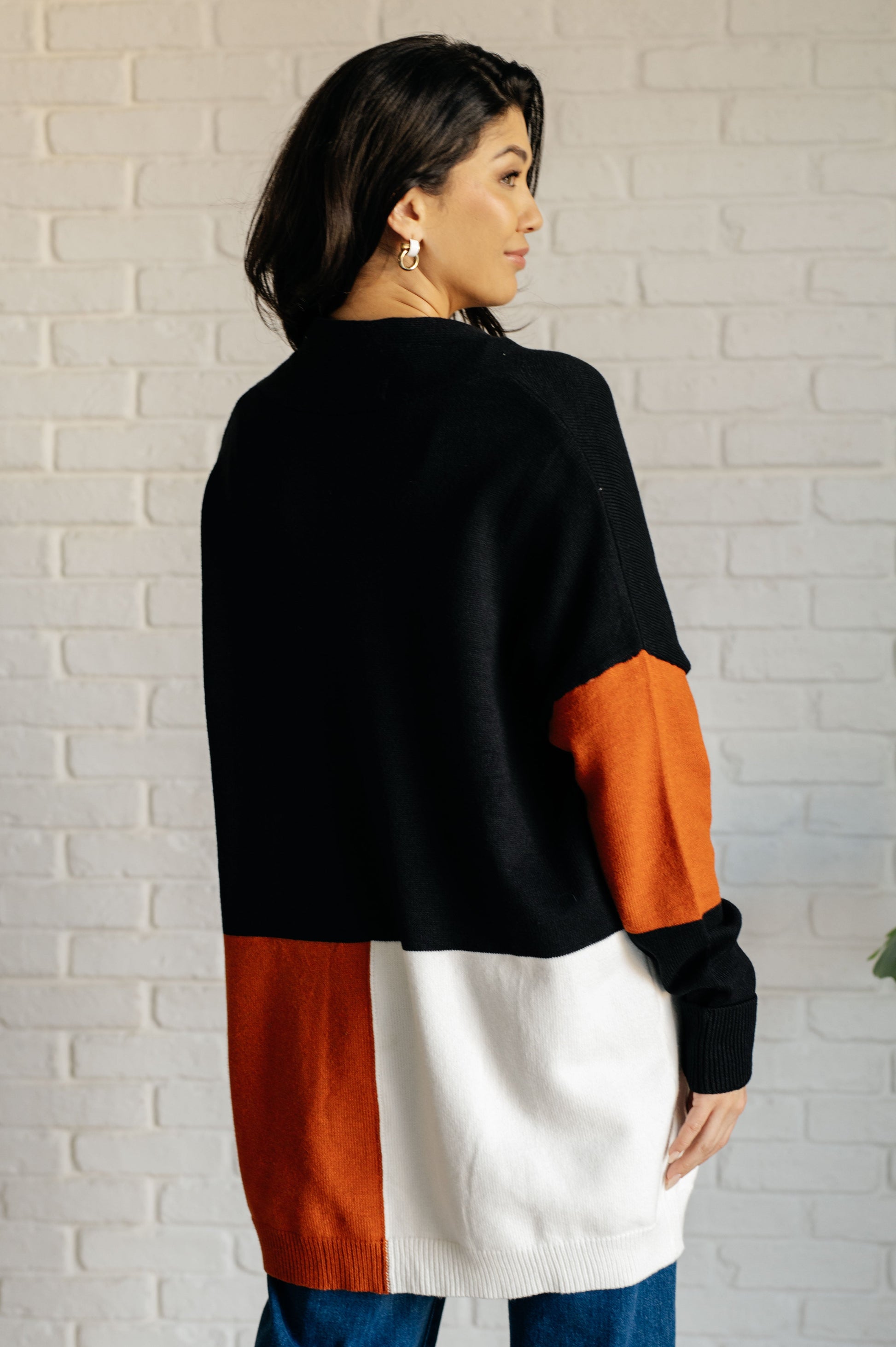 Woman wearing a black, orange, and white color-blocked sweater against a white brick wall.