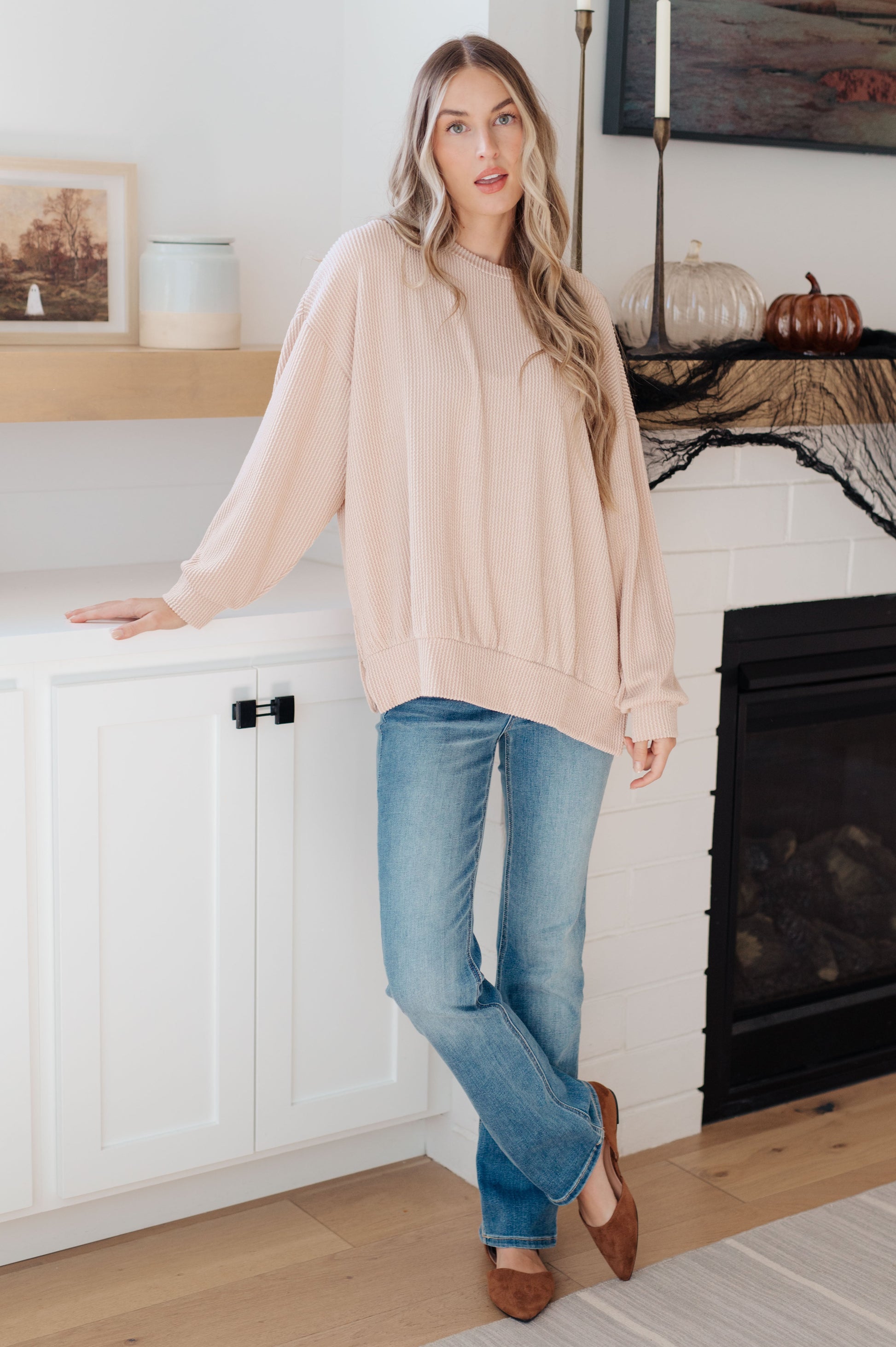 Woman wearing a beige sweater and blue jeans standing in a room with a fireplace.