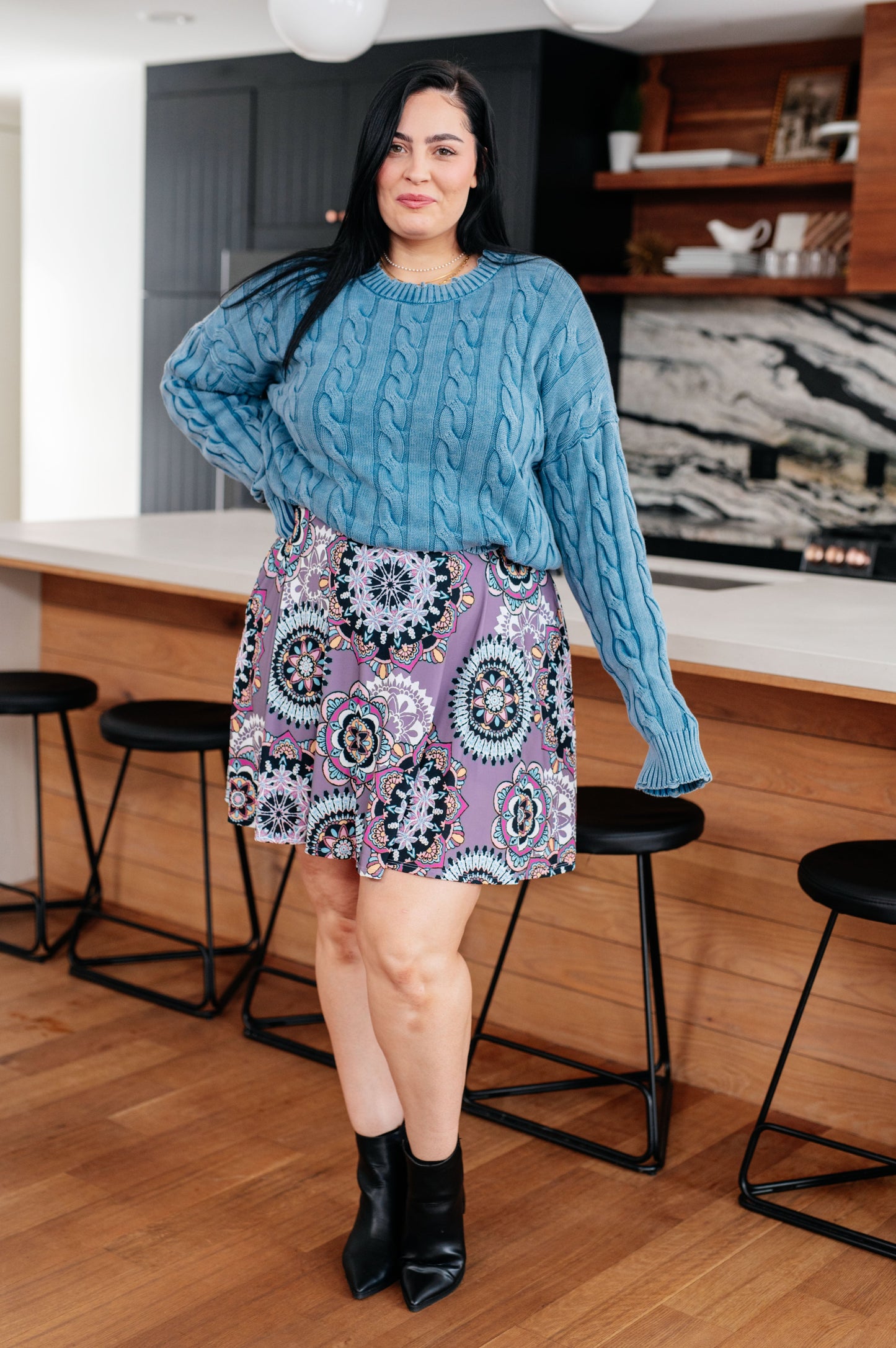 Woman wearing a blue cable knit sweater and patterned skirt in a kitchen.