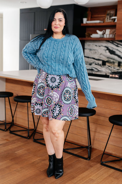 Woman wearing a blue cable knit sweater and patterned skirt in a kitchen.