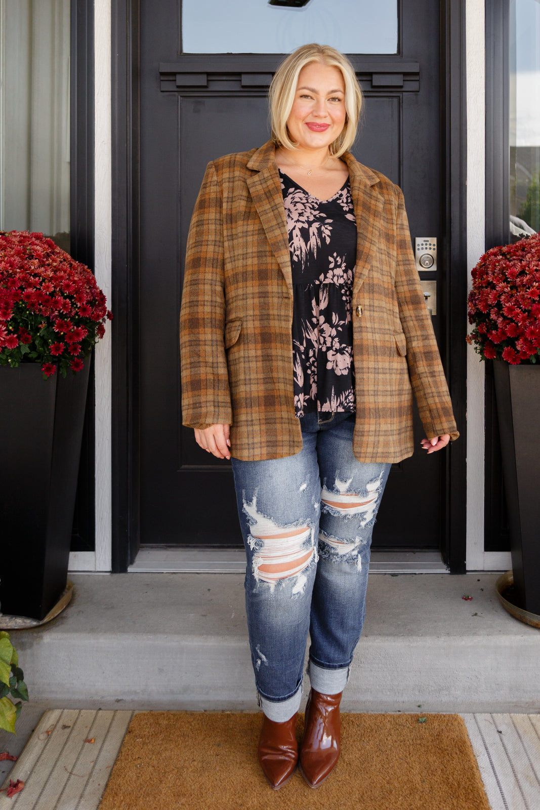 Woman standing outside a house wearing a plaid jacket, floral top, and ripped jeans.