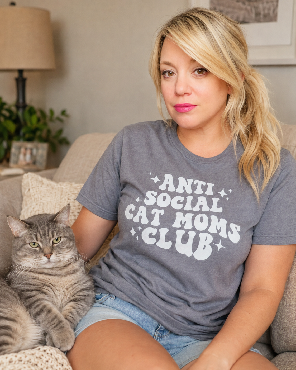 A woman sitting on a couch wearing a gray t-shirt with 'ANTI SOCIAL CAT MOMS CLUB' printed in white, next to a gray tabby cat in a cozy living room setting.