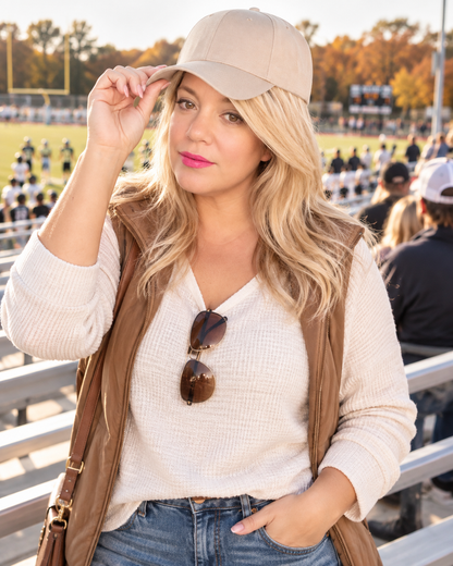 A woman at an outdoor sports event wearing a beige baseball cap, a cream ribbed knit sweater vest over a white long-sleeve shirt, and blue jeans, with sunglasses hanging from her neckline.