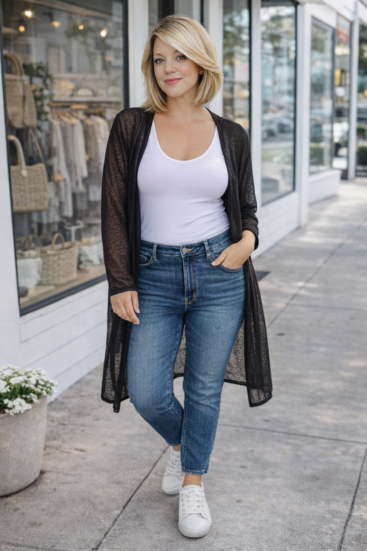 Woman wearing a white tank top, black cardigan, blue jeans, and white sneakers on a sidewalk.