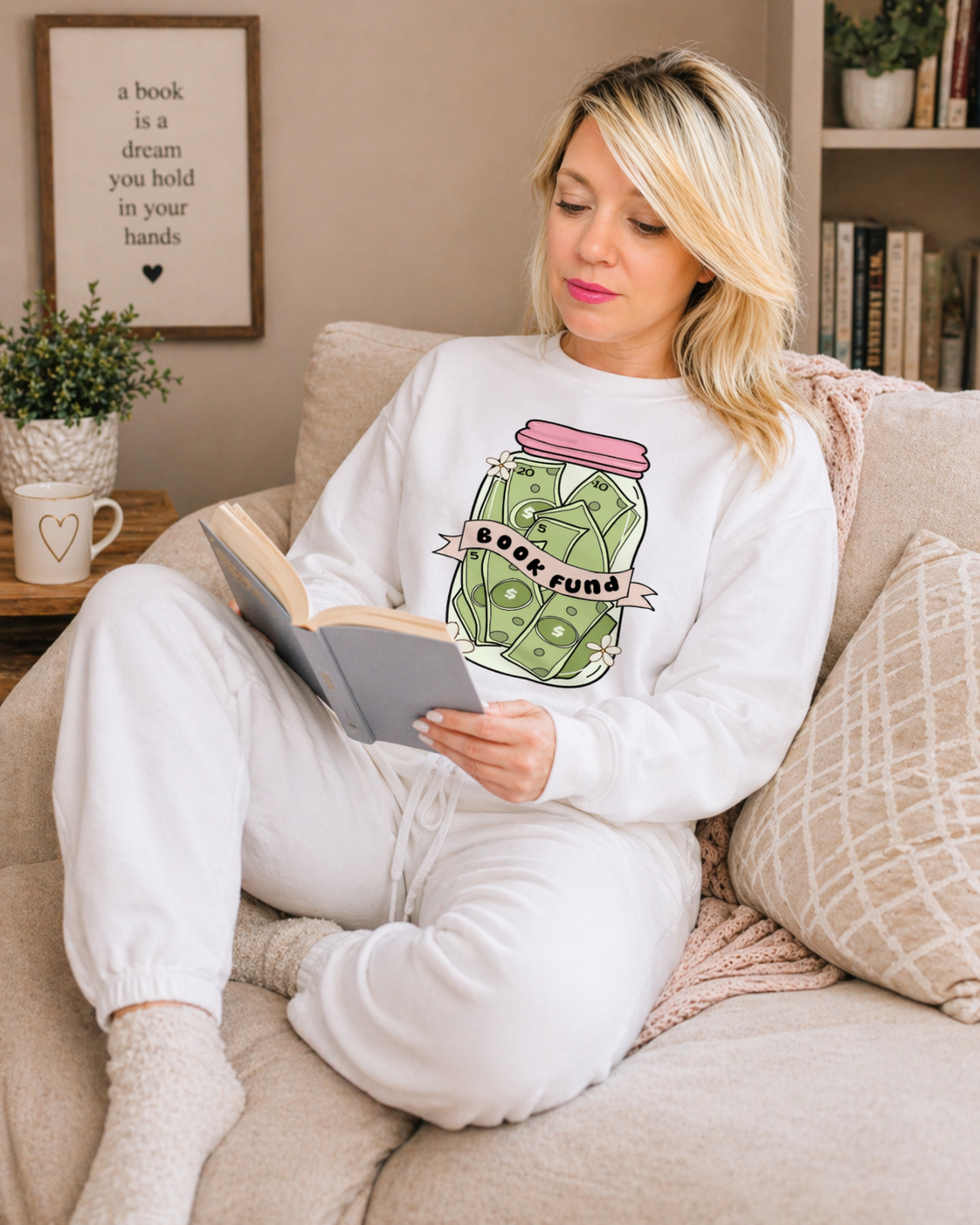 Woman reading a book wearing a white sweatshirt with a green jar design in a cozy living room.