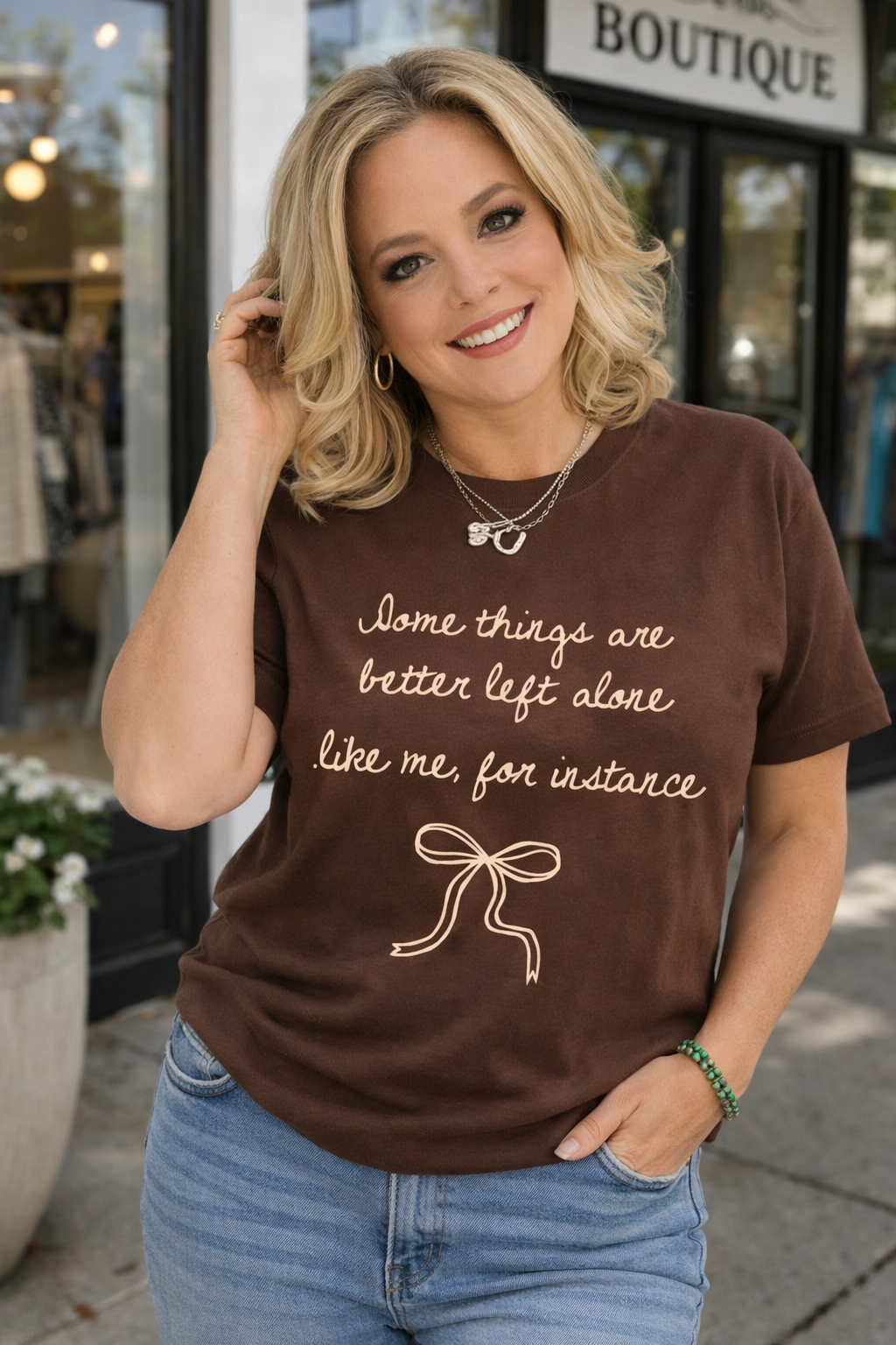Woman wearing a brown t-shirt with text and a bow graphic, standing in front of a boutique.