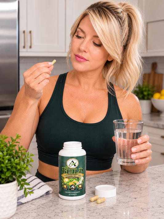 A woman in a dark green sports bra stands in a kitchen holding a glass of water and looking at a yellow supplement capsule she is holding in her other hand. In front of her is a white bottle of 'BUILT' brand herbal supplement with its cap off, next to two yellow capsules.