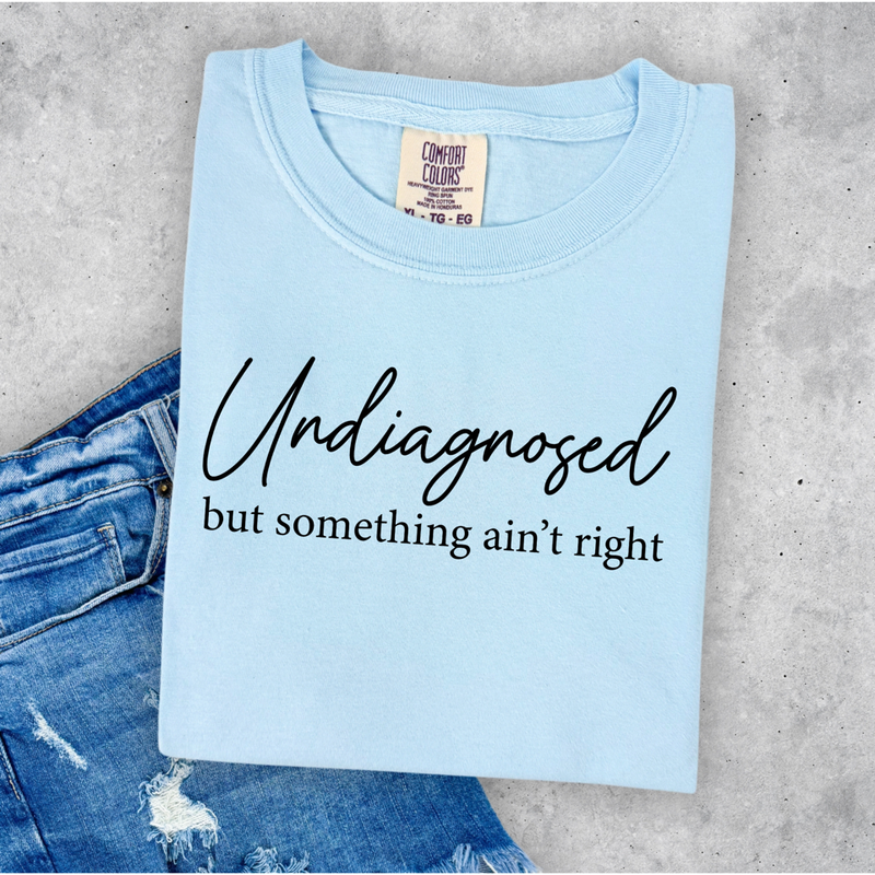 Light blue t-shirt with 'undiagnosed but something ain't right' text on a concrete surface with jeans.