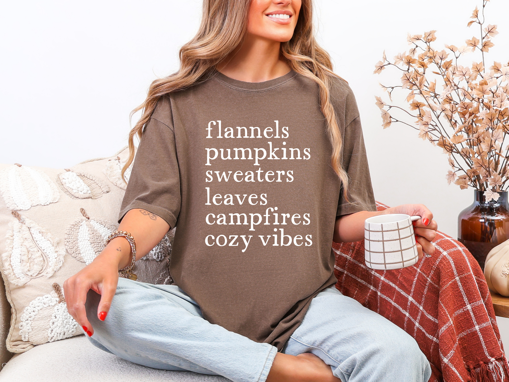 Woman wearing a brown shirt with text, sitting on a couch with a blanket and mug.