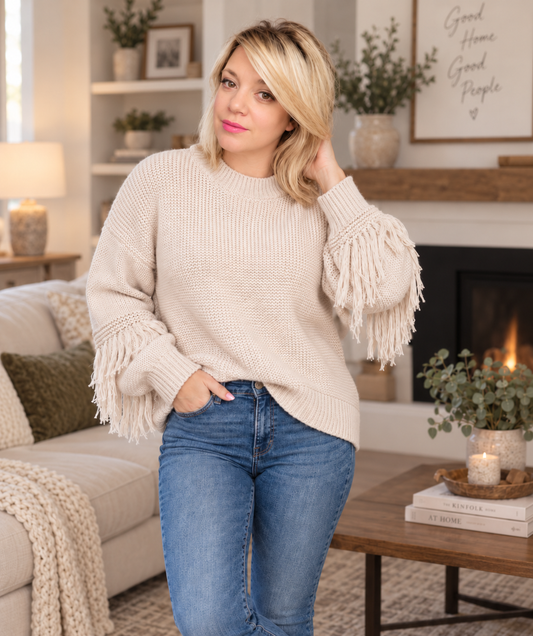 A woman wearing a cream-colored ribbed knit sweater with long fringe sleeves, paired with blue jeans, standing in a cozy living room with a fireplace and sofa in the background.