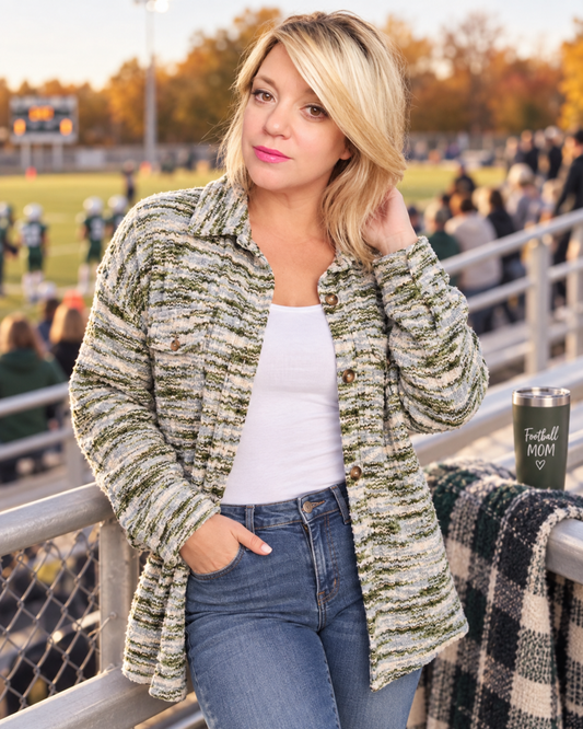A woman stands on a sports field railing at sunset wearing a textured, heathered green and beige button-up cardigan over a white tank top and blue jeans. A black insulated tumbler with 'Football Mom' printed on it rests on the railing beside her.