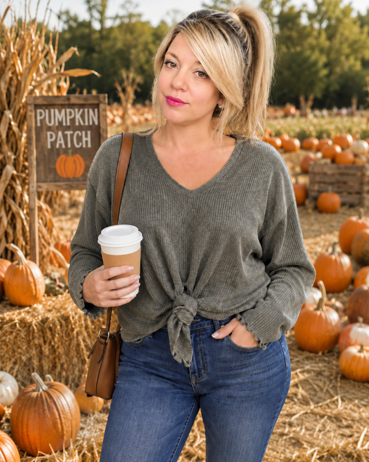 A woman stands in a pumpkin patch wearing a ribbed knit V-neck sweater tied at the waist, blue jeans, and holding a coffee cup. A 'Pumpkin Patch' sign and hay bales are visible in the background.