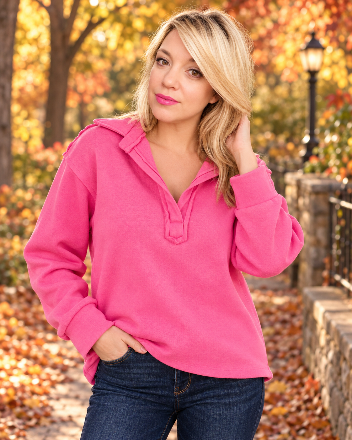 A woman wearing a bright pink long-sleeve mock neck pullover sweater and dark blue jeans, standing outdoors on a path covered with fallen leaves, with autumn trees in the background.