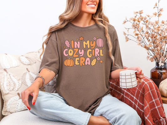 Woman wearing a brown shirt with colorful text, sitting on a couch with a mug and blanket.