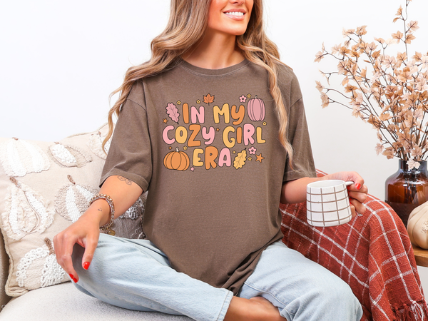 Woman wearing a brown shirt with colorful text, sitting on a couch with a mug and blanket.
