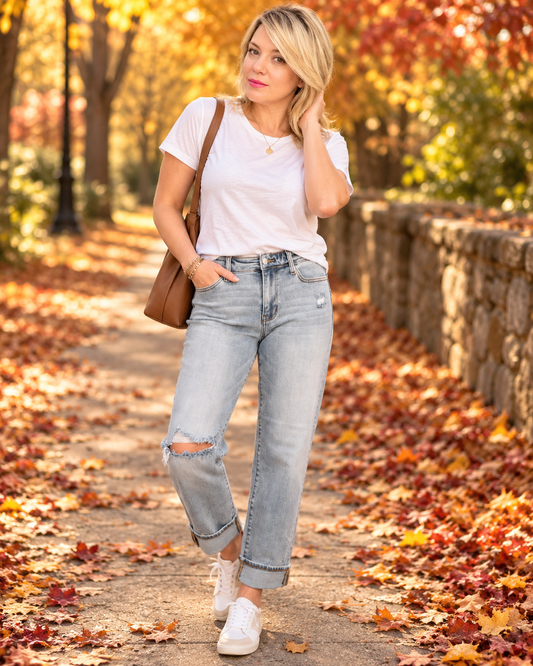 A woman stands on a leaf-covered path in an autumn park, wearing a white short-sleeve t-shirt, light-wash distressed jeans, white sneakers, and carrying a brown shoulder bag.