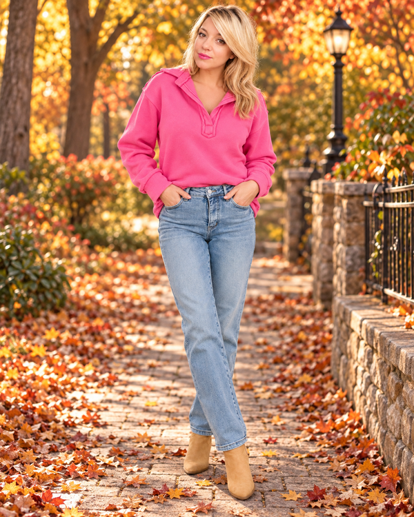 A woman stands on a stone path covered with fallen autumn leaves, wearing a bright pink long-sleeve polo-style top, light blue jeans, and tan ankle boots. Trees and shrubs with orange and yellow foliage are in the background.