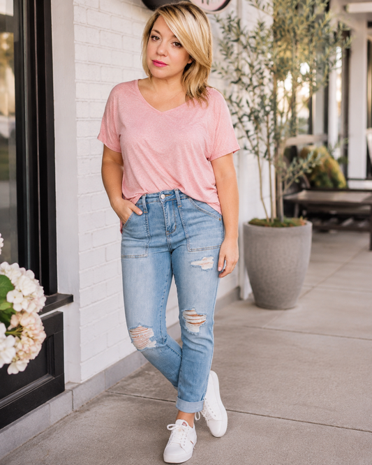 A woman posing on a sidewalk wearing a light pink v-neck t-shirt, light-wash distressed blue jeans, and white sneakers.