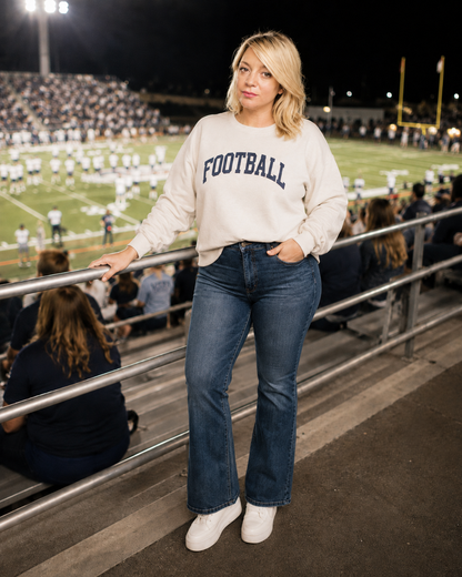 A woman stands in a stadium seating area at night, leaning on a metal railing. She is wearing a cream sweatshirt with 'FOOTBALL' printed in navy blue arched letters across the chest, paired with blue jeans and white sneakers.