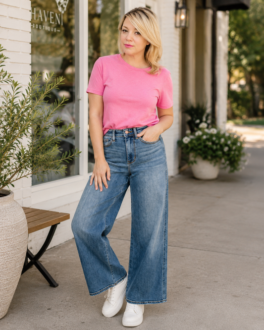 Woman wearing a pink shirt and blue jeans standing outdoors near a building.